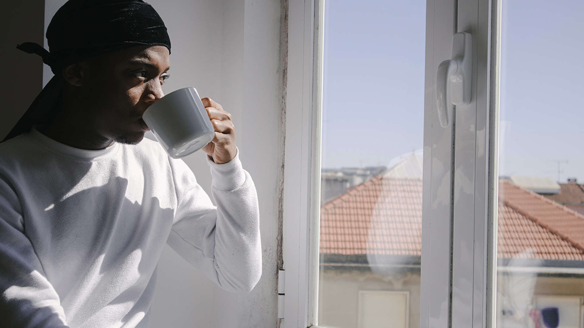 A young man looking out the window with a white long sleeve top on, drinking out of a mug