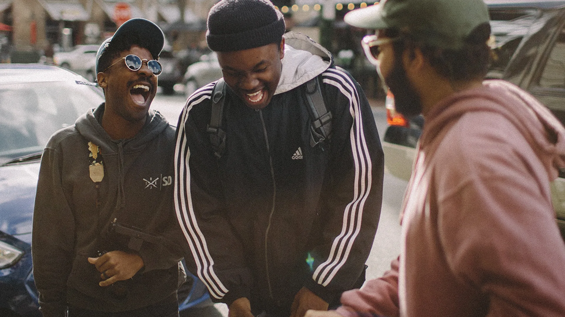 Three young men laughing and leaning against car on the street.