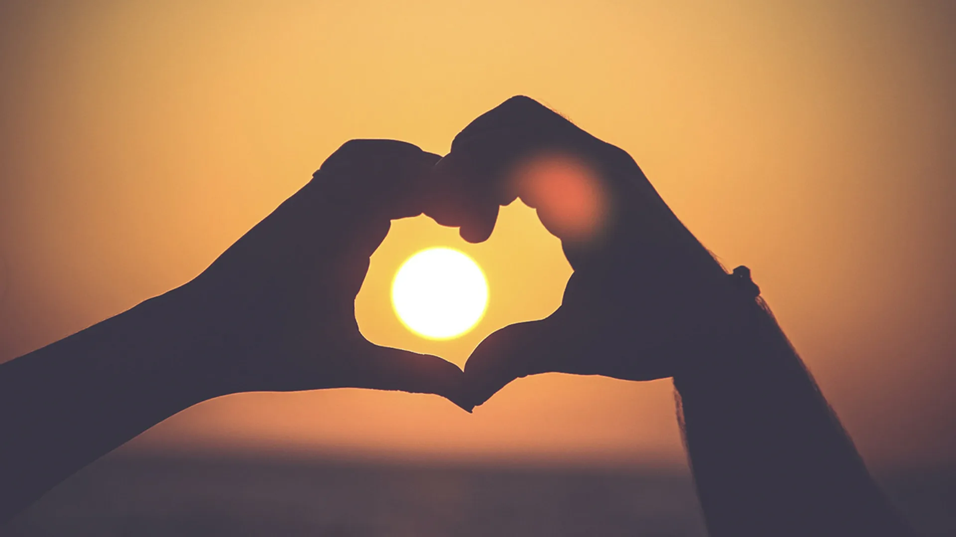 The silhouette of two people's hands forming heart at sunset, with the sun positioned inside the heart.