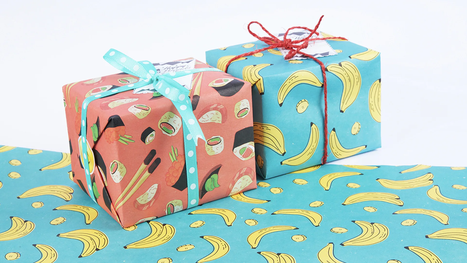 Two wrapped presents on white background. One is wrapped in blue paper with banana pattern, and another one is wrapped in red paper with sushi pattern.