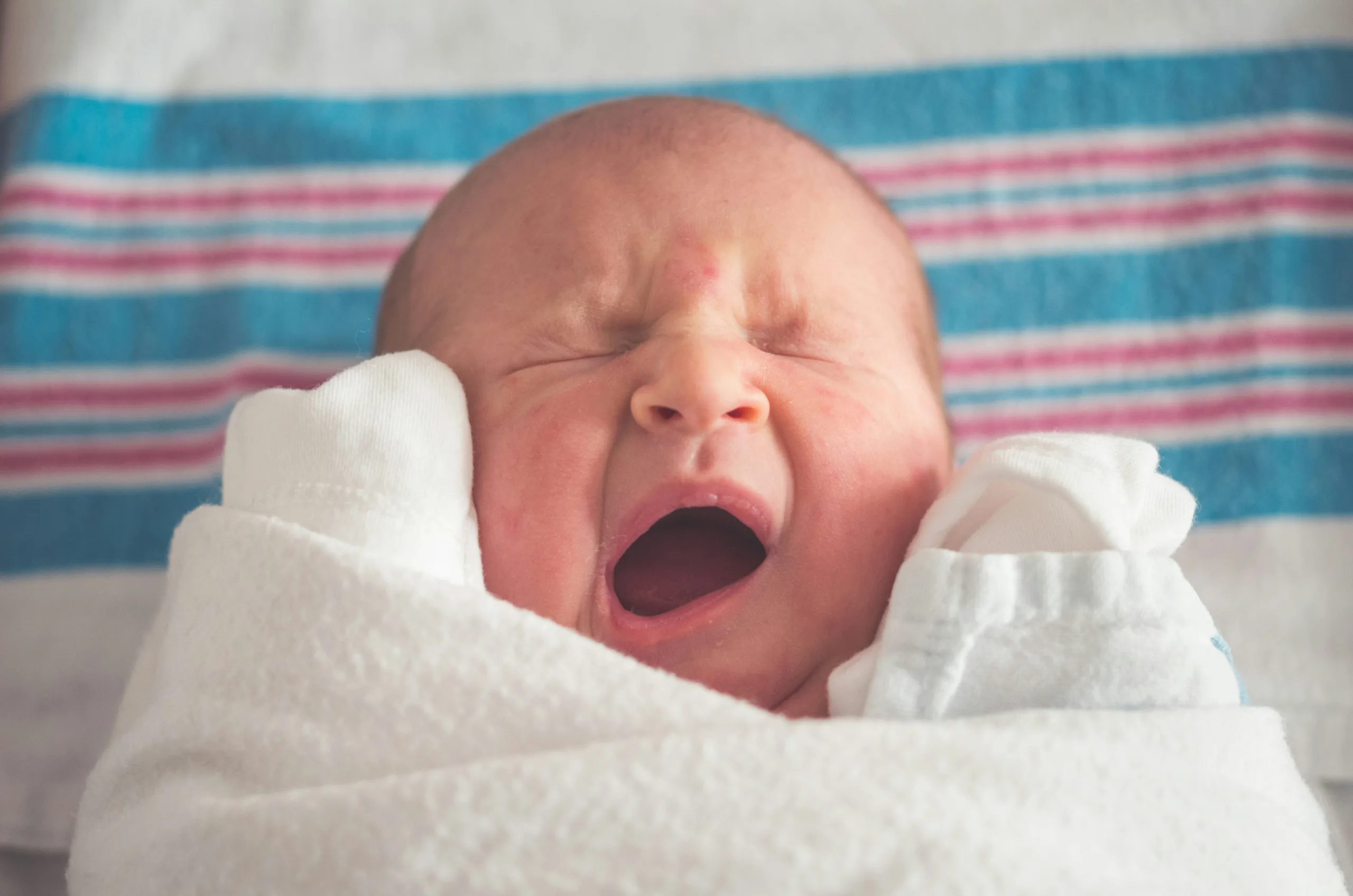Photograph of a baby crying swaddled in a white blanket lying on a striped cloth