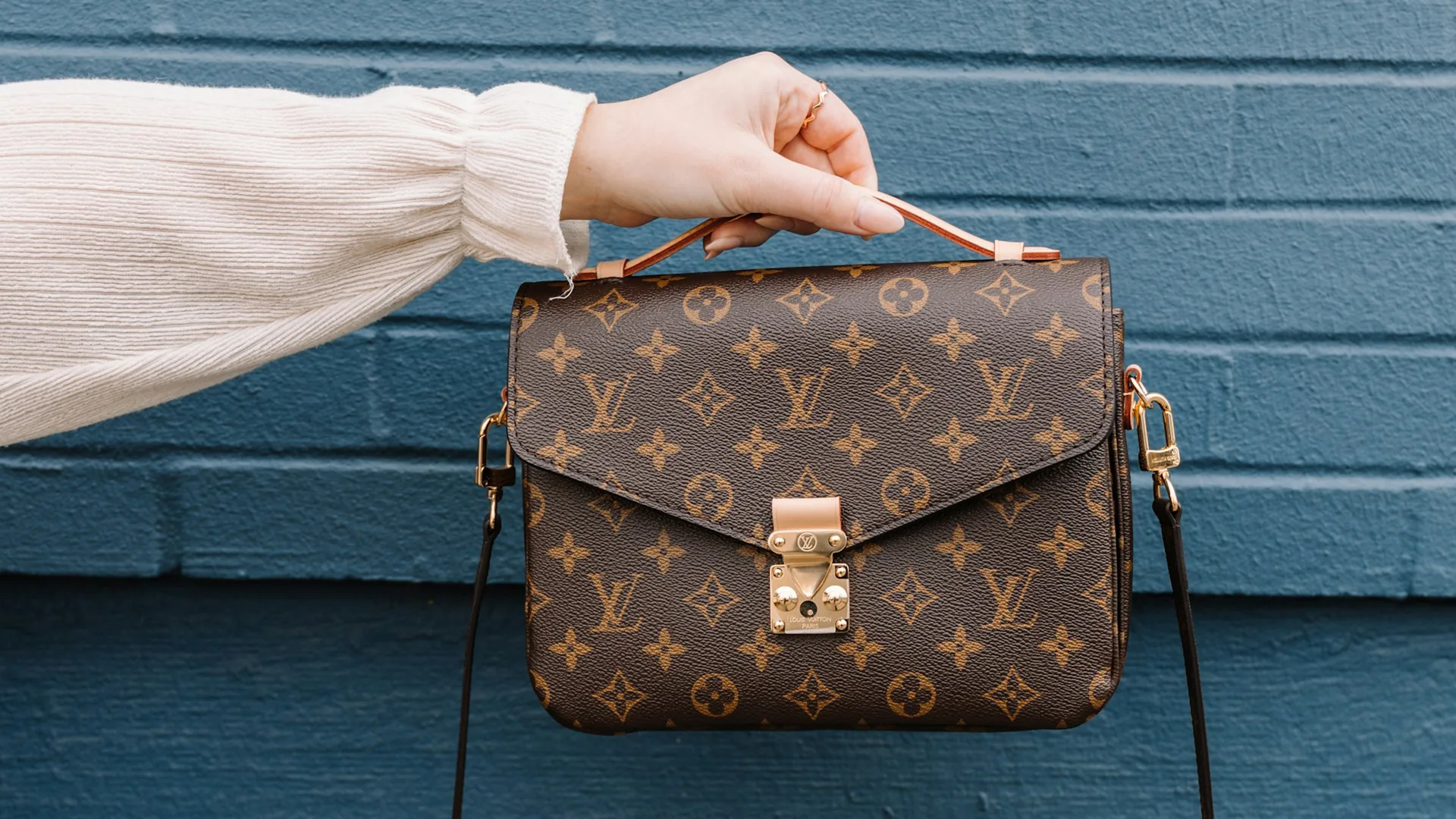 Persons hand holding a Louis Vuitton handbag against a blue brick wall