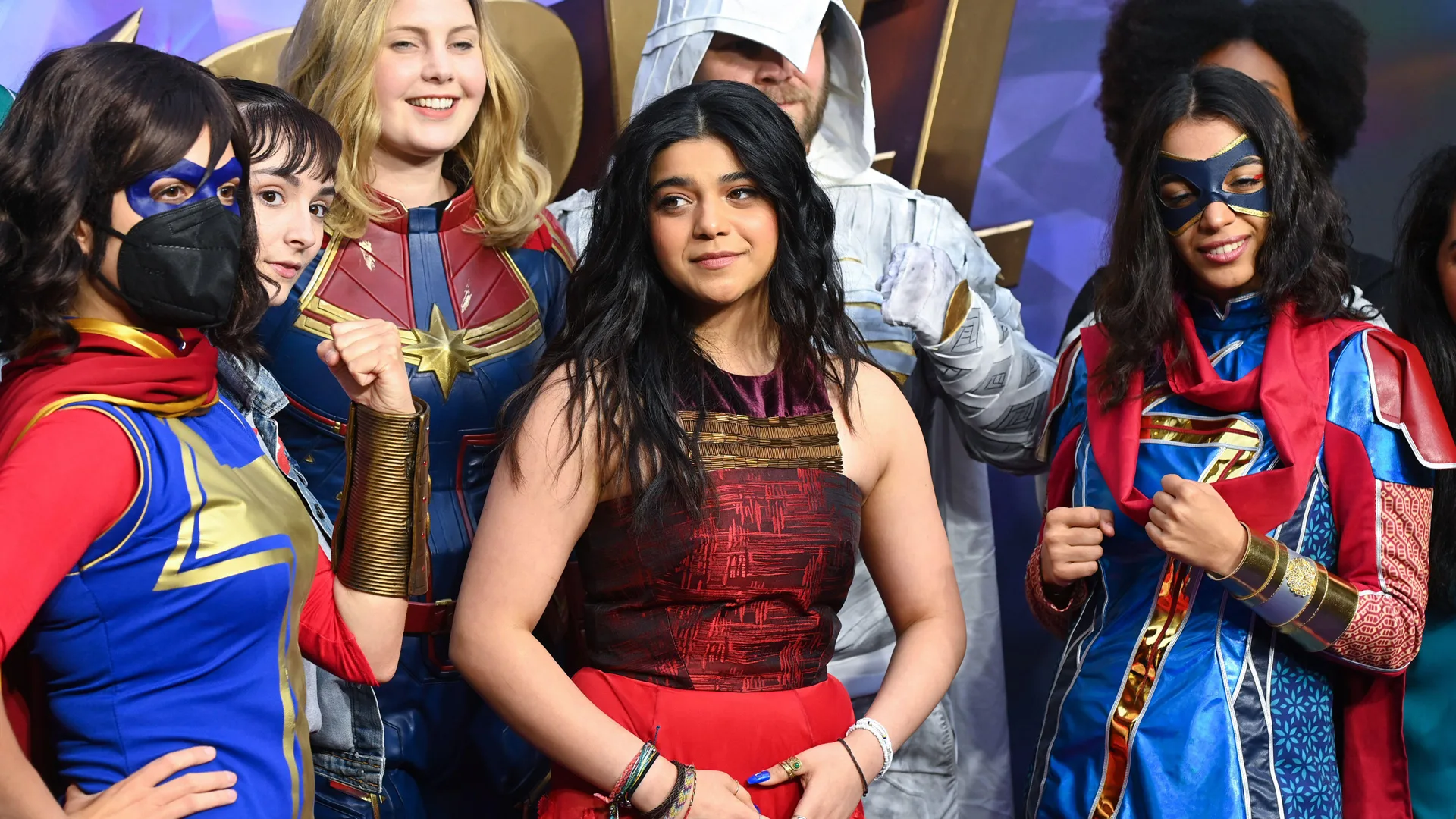 A photograph of Iman Vellani attending the Ms Marvel premiere wearing a black and red dress surrounded by other cast members in superhero outfits