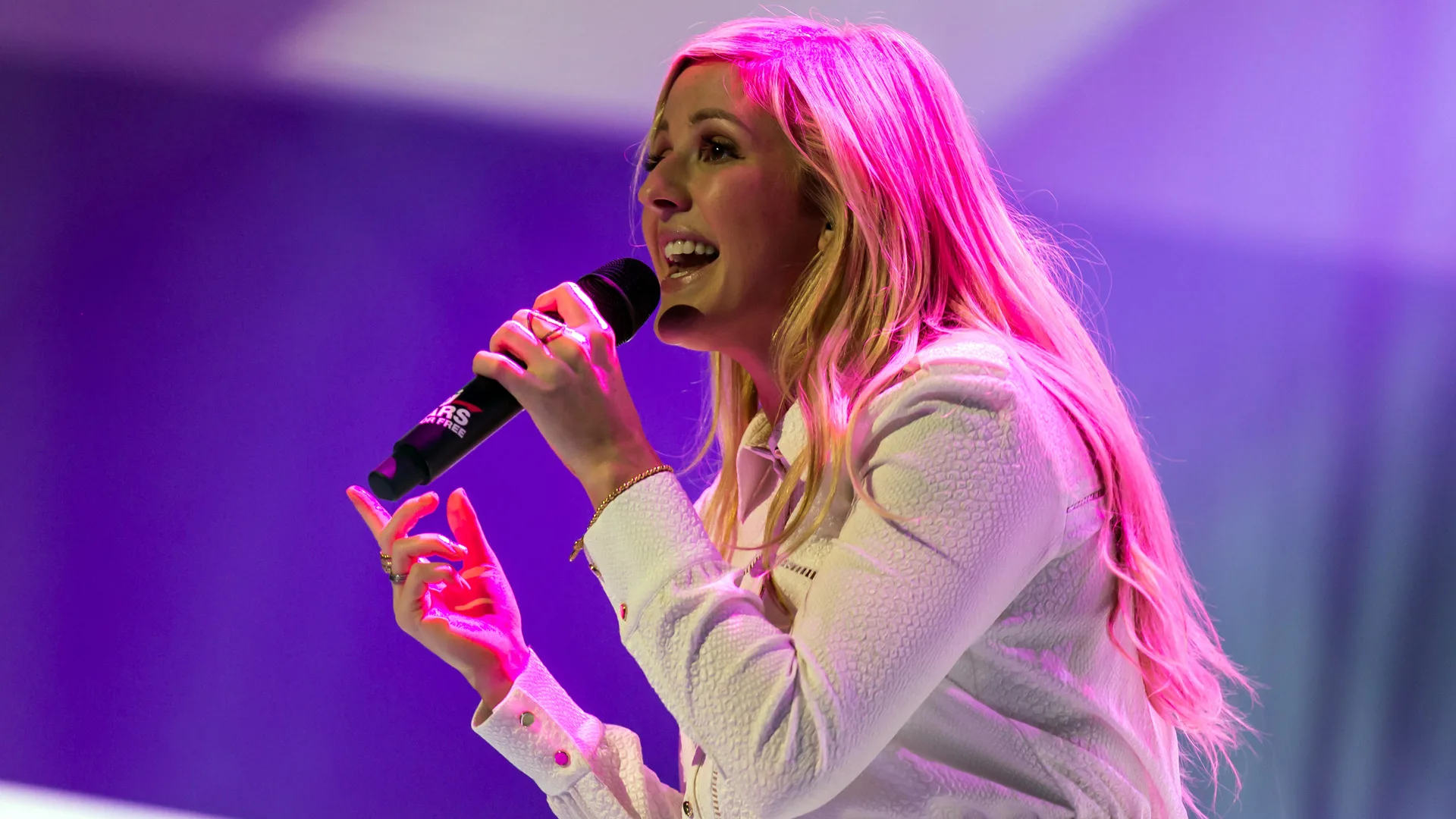 A photograph of Ellie Goulding singing on stage with a microphone. She has her long blonde hair down and is wearing a white top. The background is lit up purple.