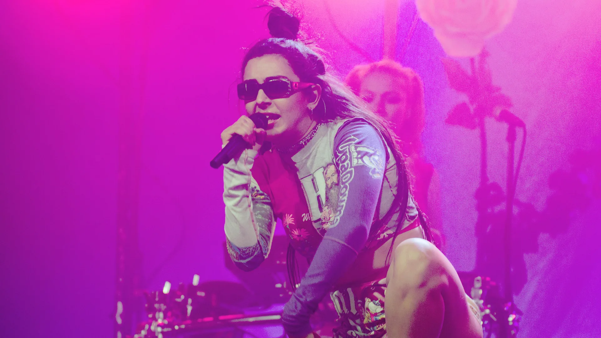 A photo of the singer Charlie XCX on stage singing with a mic. She is wearing sunglasses and a cropped top - her hair is up in a top knot. The background is pink and the drummer is just out of focus.