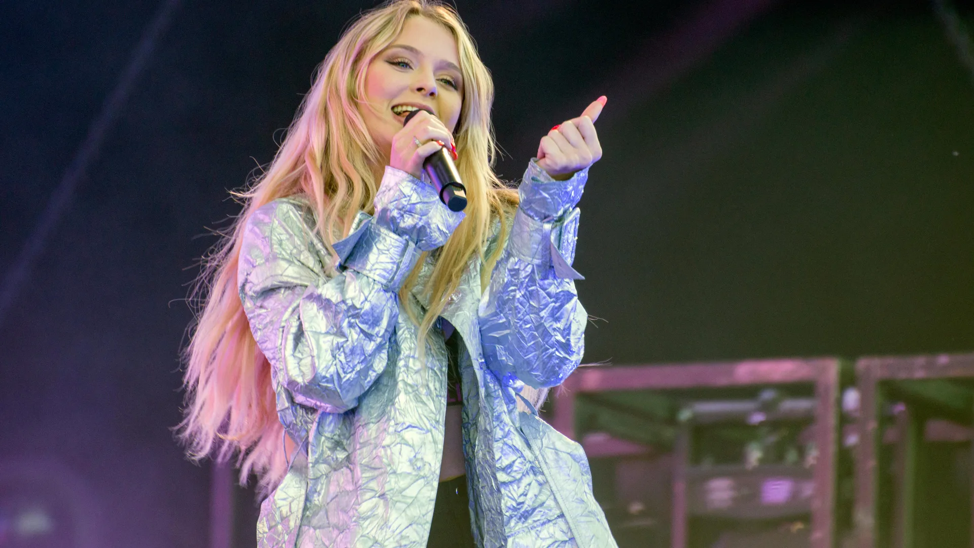 A photograph of Zara Larsson performing on stage with a microphone. She is wearing a silver foil jacket and her hair is long and blonde, against a dark stage background.
