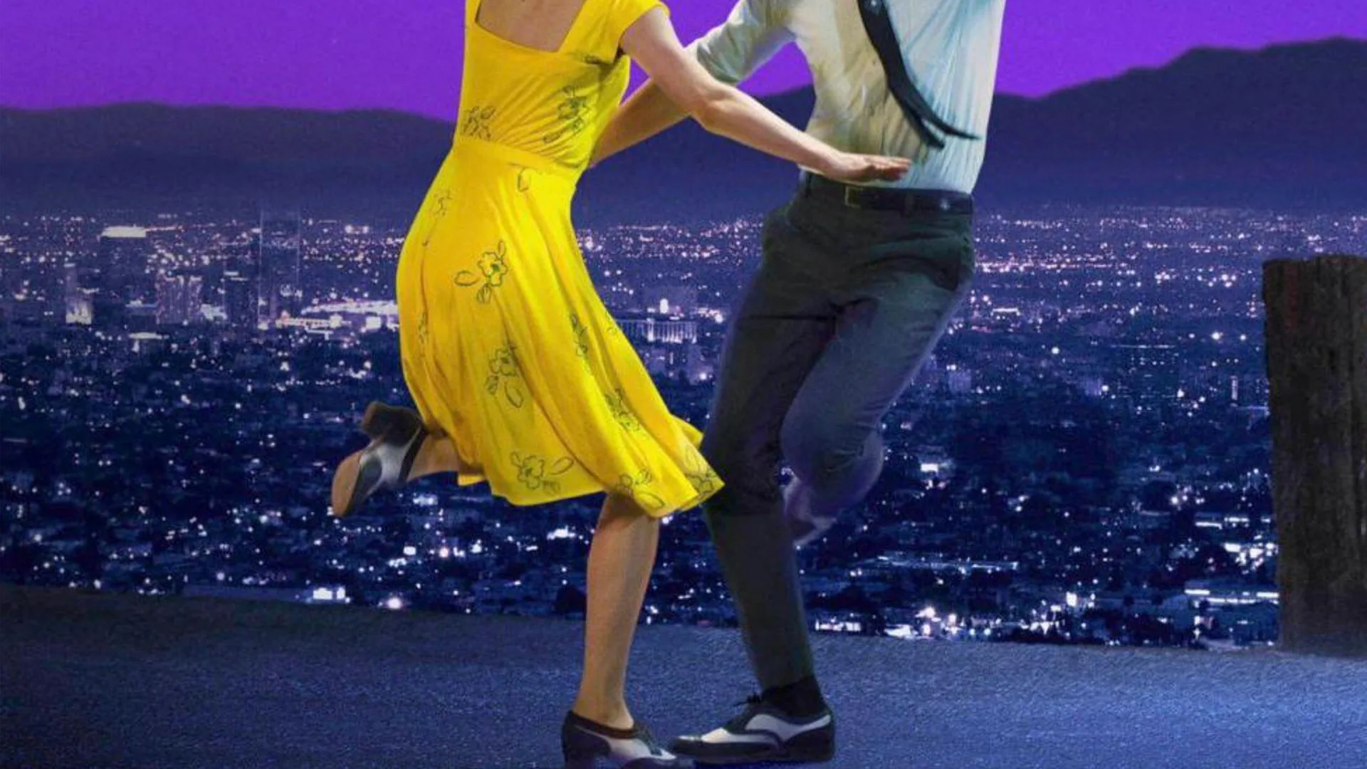 A photograph of two people dancing. The woman is wearing a yellow dress and the man is wearing a suit. They are dancing against a city at night background.