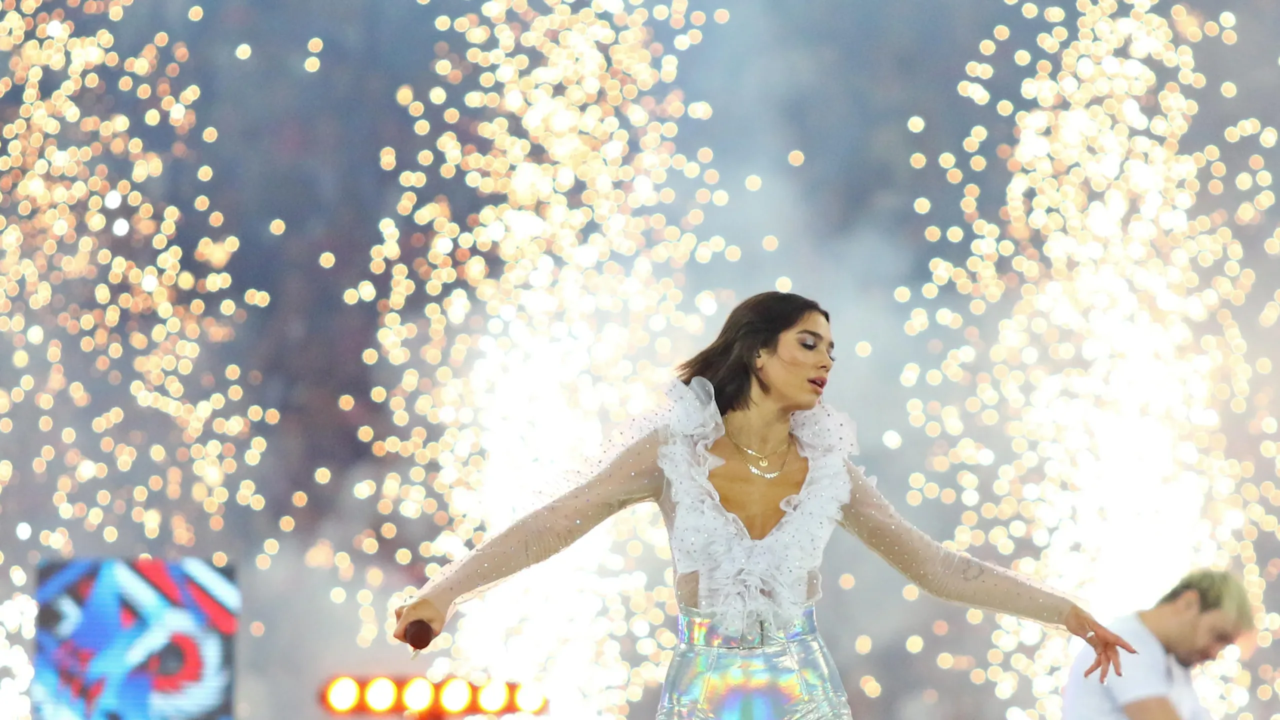 Dua Lipa performs onstage in white outfit with fireworks behind her