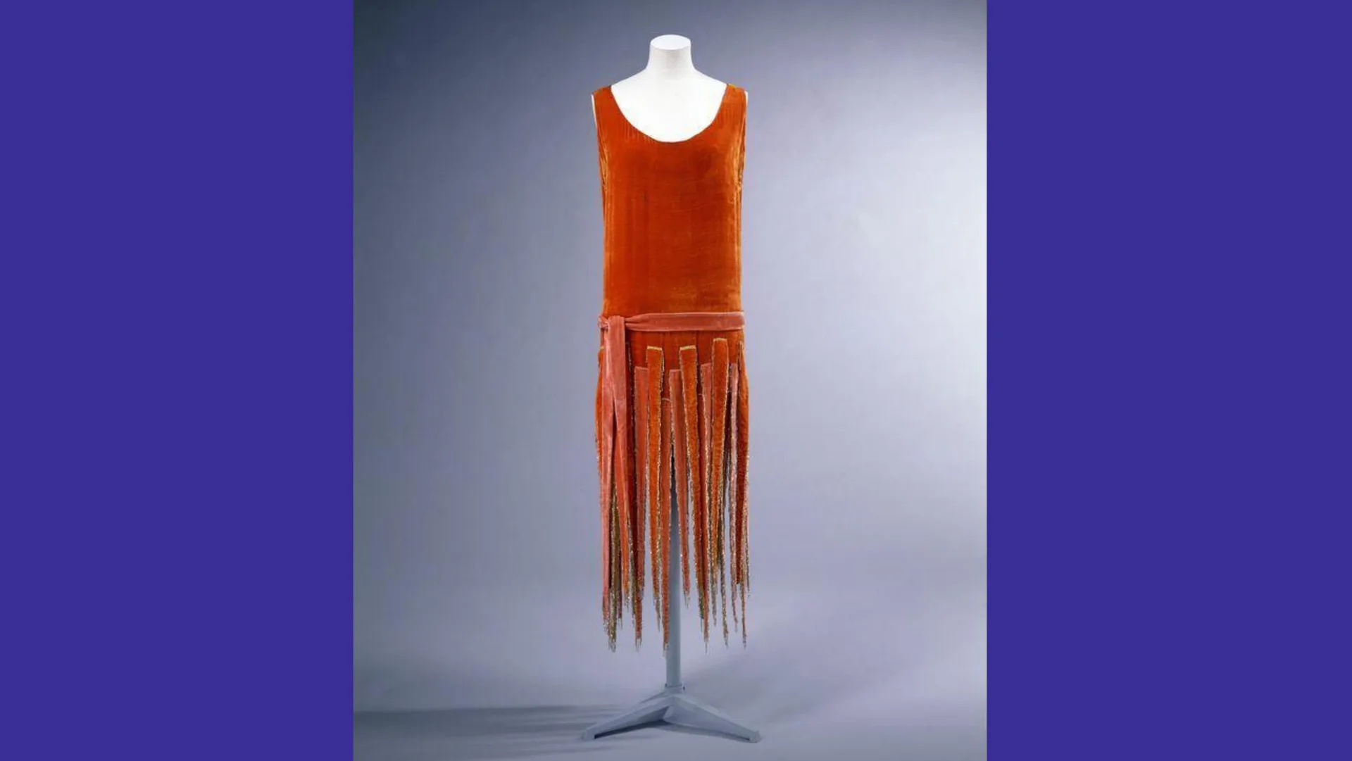 A photograph of a burnt orange silk velvet dress with tassles hanging down made of the same material, against a grey background with blue borders