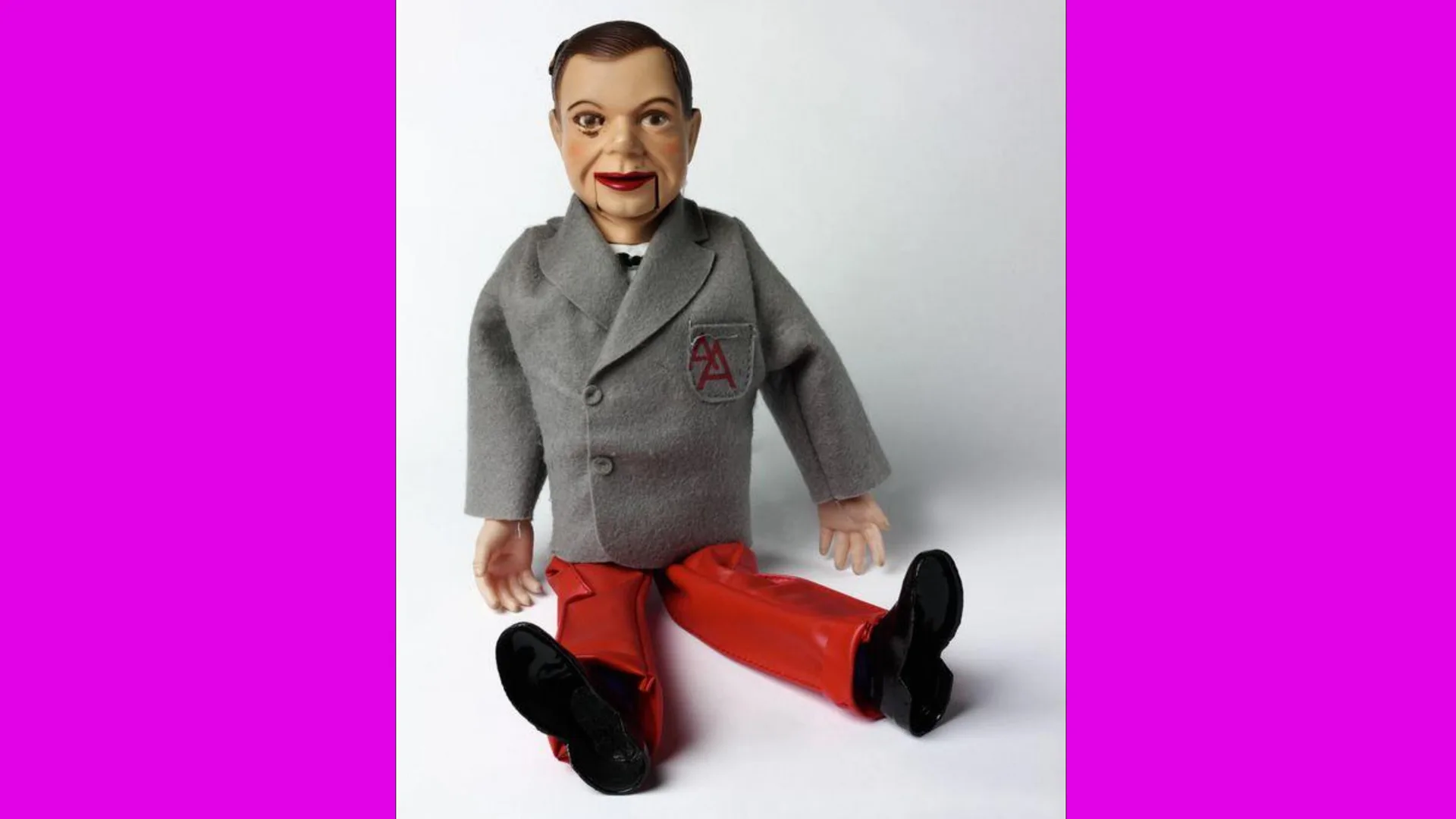 A photo of a ventriloquist doll wearing a grey jacket and red trousers in a seated upright position on a grey background with pink borders