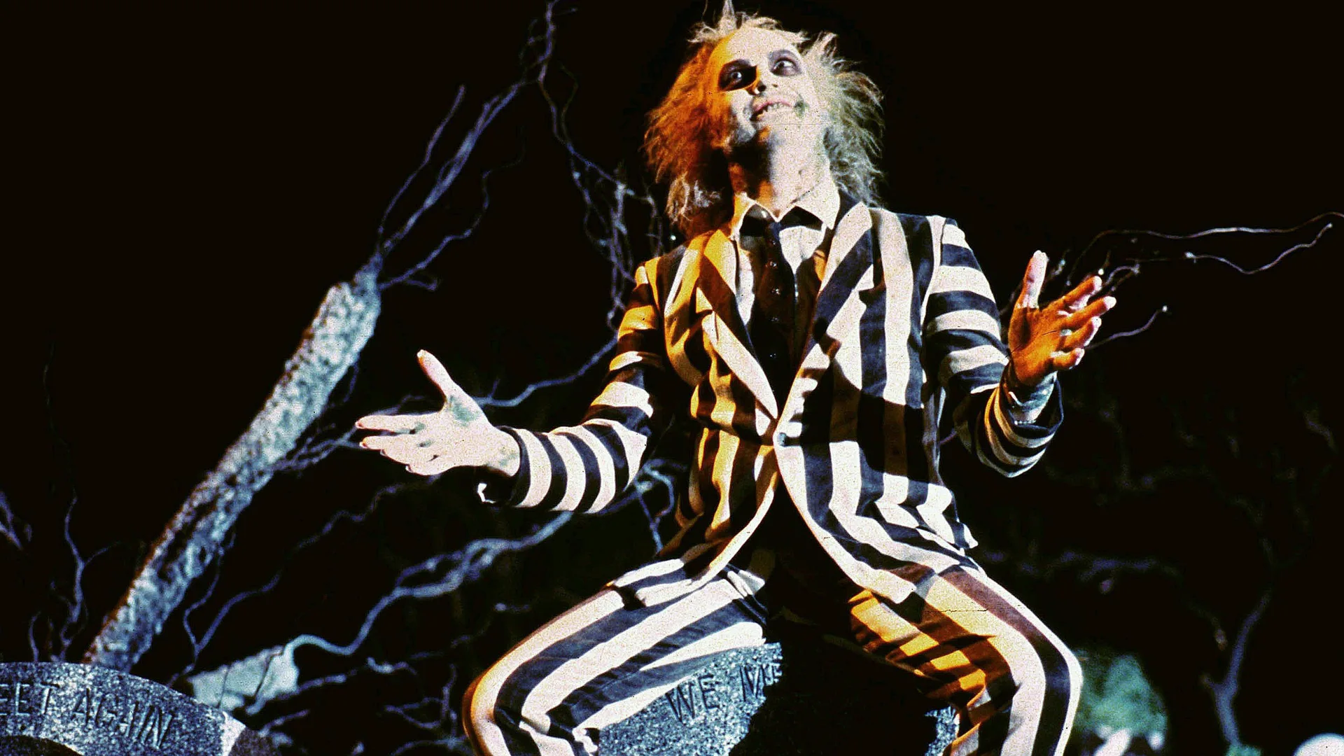 A still from the movie Beetlejuice showing Michael Keaton as the character Beetlejuice wearing his iconic black and white striped suit sat on a gravestone with his arms stretched out and a grin on his face against a dark cemetary background.