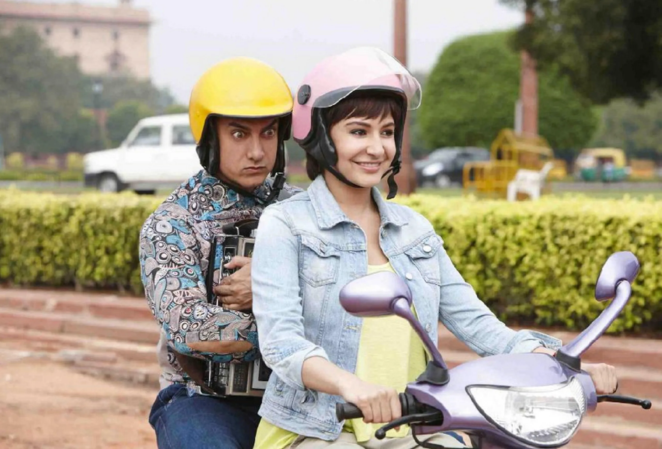 A still of Aamir Khan riding on a motorbike looking worried clutching books with a yellow helmet and driving it is Anushka Sharma who is smiling wearing a pink helmet