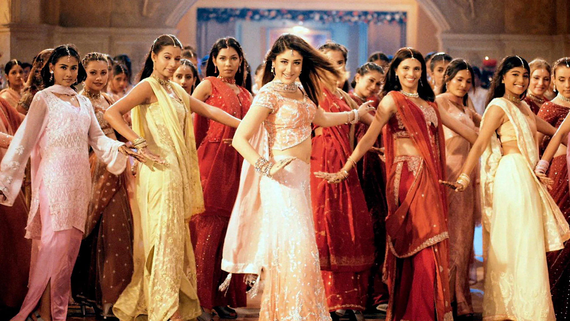 A photograph of many women dancing wearing colourful traditional indian outfits with hands splayed out smiling looking directly at the viewer