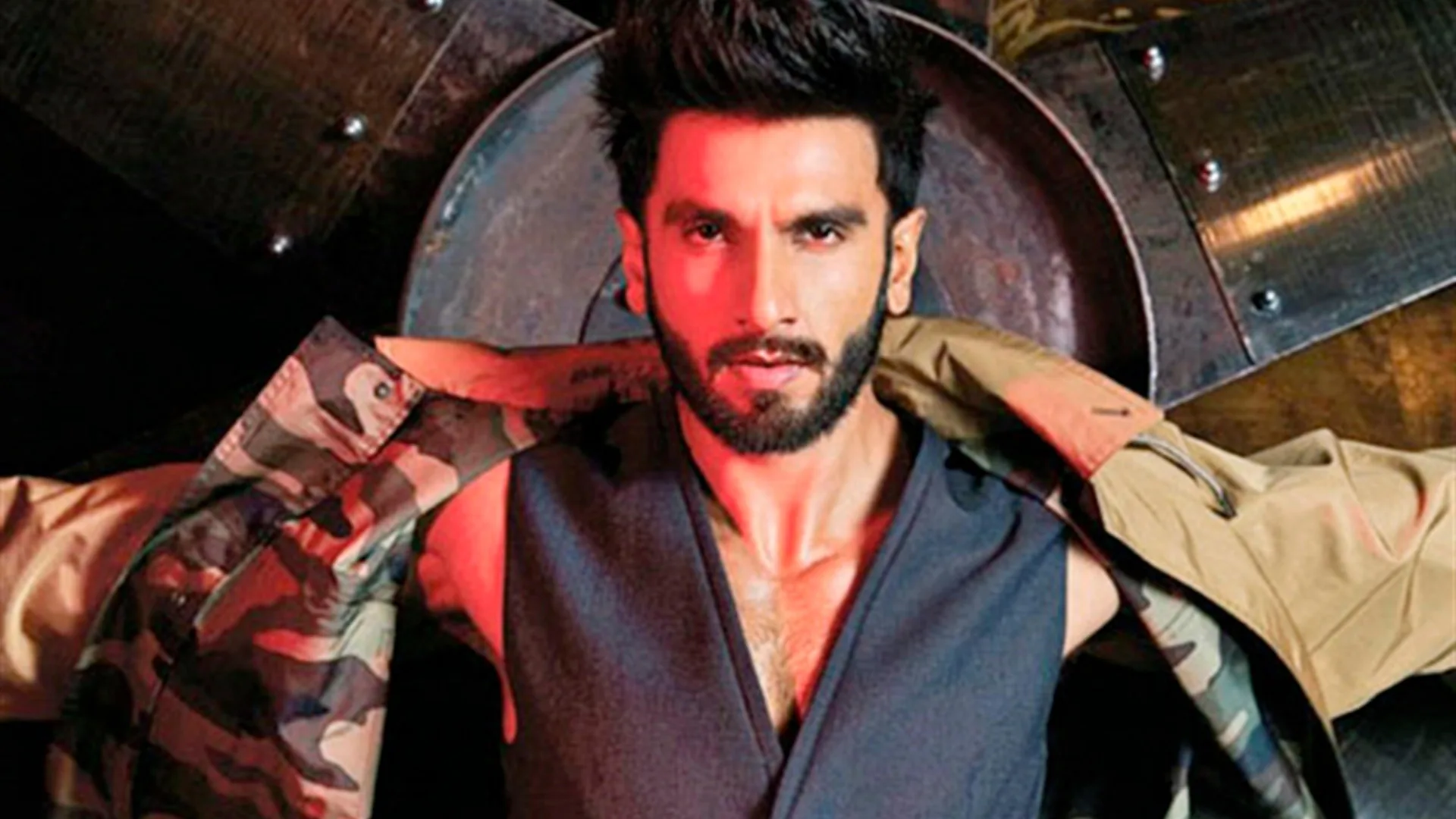 A photo of the actor Ranveer Singh wearing a black shirt midway putting a beige coat with army camoflage lining against a black and grey background