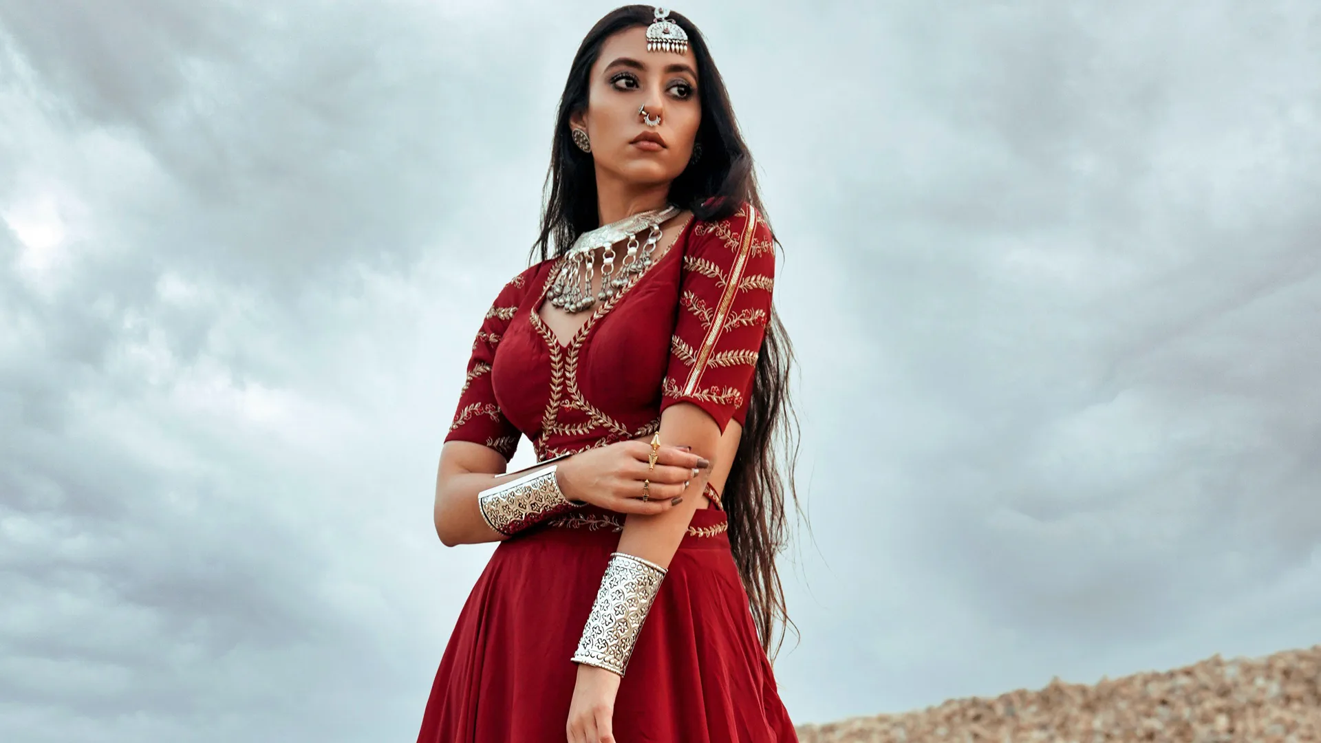 A photo of a woman wearing a red lehenga holding her arm looking out to the distance against a grey cloudy sky