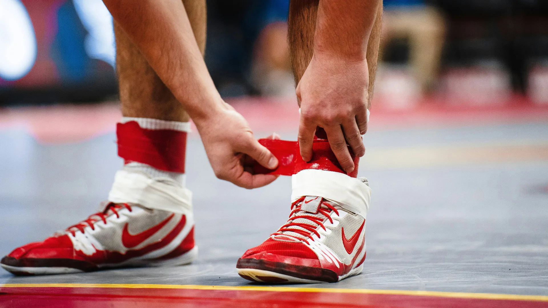 A photo of a wrestler tieing up their red and white shoes stood on a grey floor