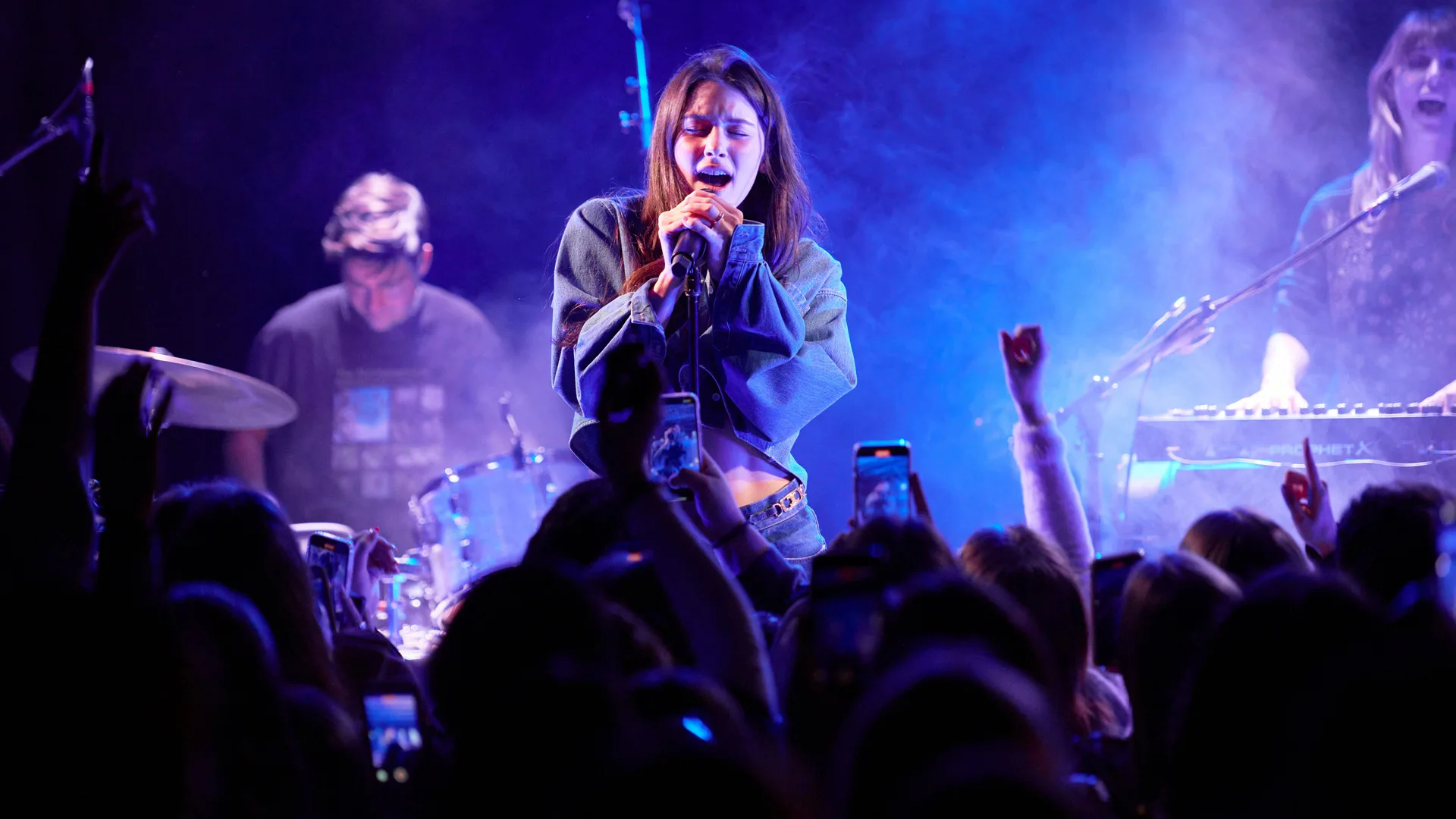 A photo of singer Gracie Abrams performing on stage singing into a mic with purple lights around her as the crowd cheers on.