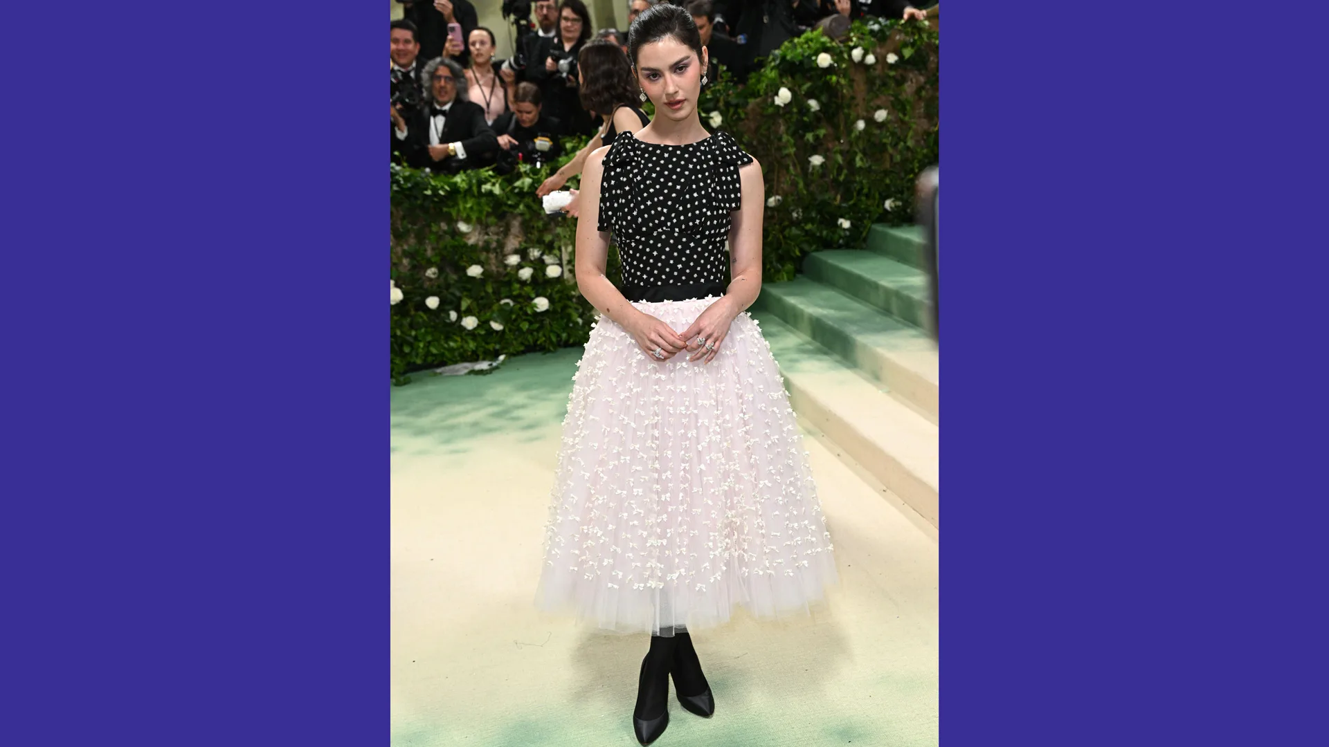 A photo of Gracie Abrams wearing a black top and white skirt on the Met Gala 2024 with purple borders to the image