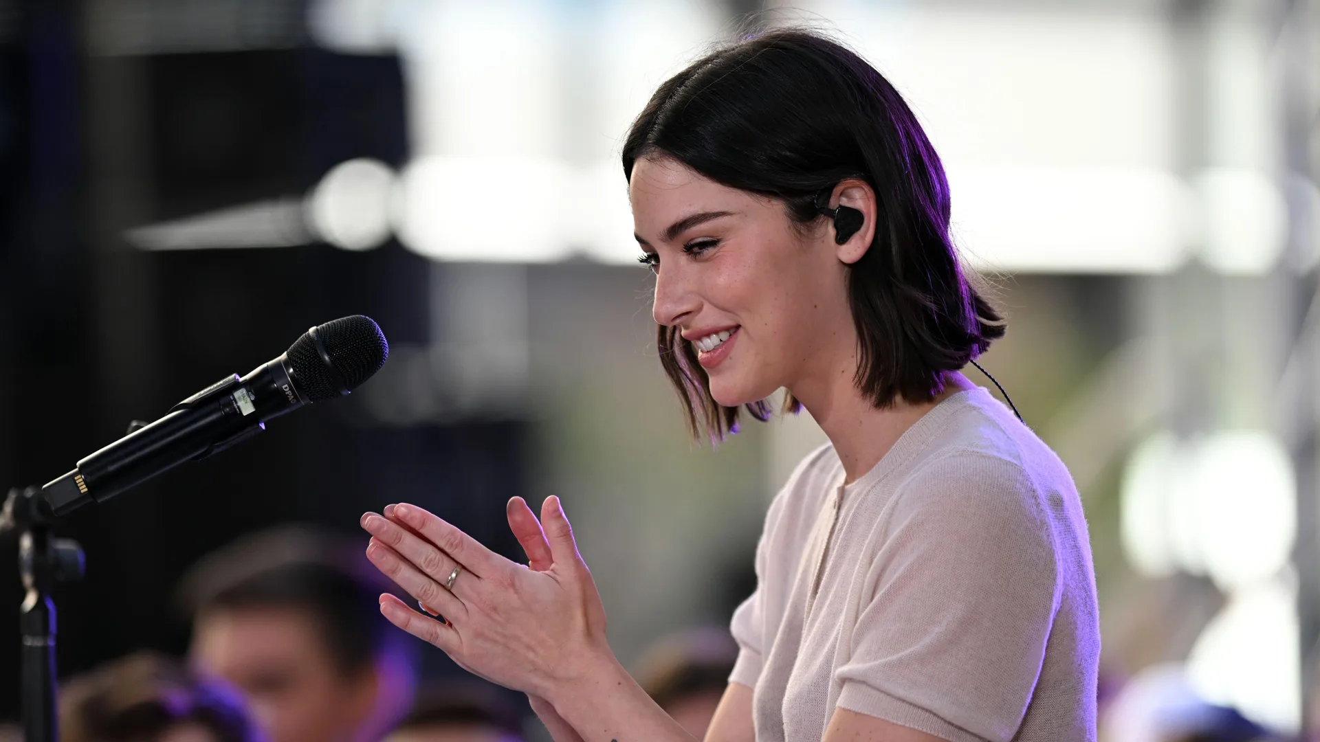 A photo of Gracie Abrams sat with her hands together smiling with a mic in front of her.