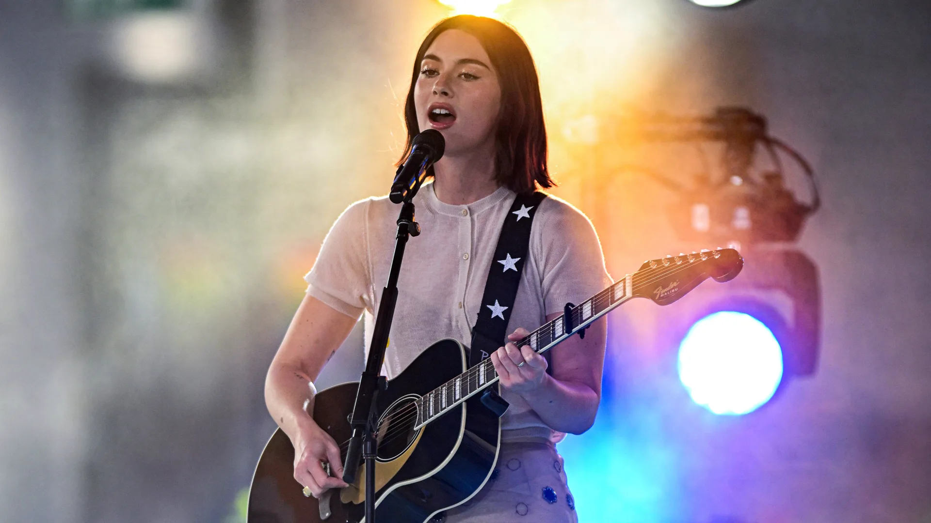 A photo of the singer Gracie Abrams performing on stage with her guitar and blue and gold lights and smoke around her