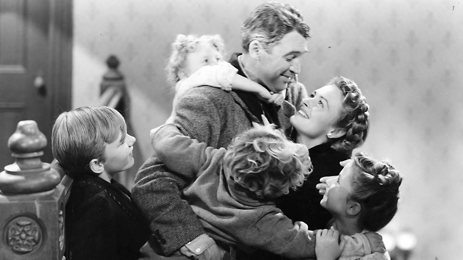 A black and white photo of the actor Jimmy Stewart playing George Bailey with his wife and children embracing him and laughing in a livingroom setting.